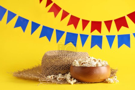 Schale mit leckerem Popcorn, Strohhut und Fahnen für die Festa Junina auf gelbem Hintergrund
