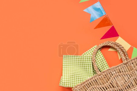 Stylish wicker bag with napkin and flags for Festa Junina celebration on color background