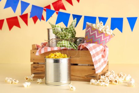 Wooden basket with tasty popcorn, canned corn and flags for Festa Junina celebration on color background