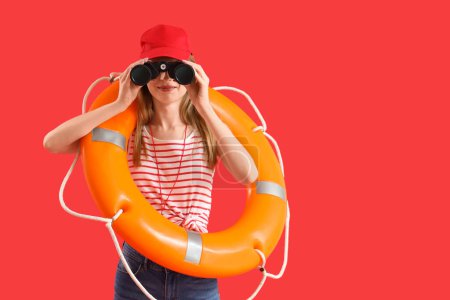 Young female lifeguard in cap with rescue ring looking in binocular on red background