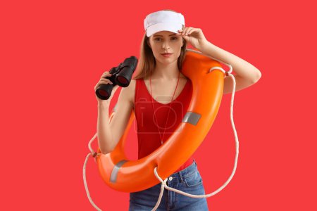 Young female lifeguard in cap with rescue ring and binocular on red background