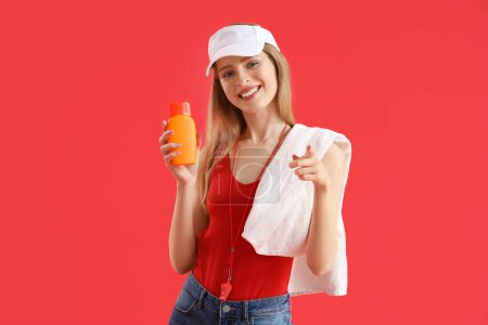 Young female lifeguard in cap with towel and sunscreen on red background