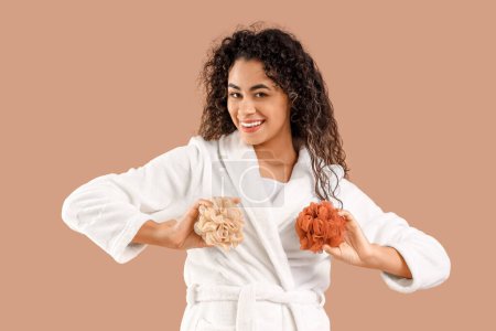 Young African-American woman in bathrobe with loofahs on brown background