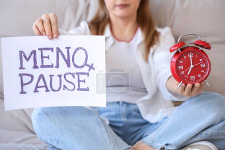 Mujer madura sosteniendo papel con palabra MENOPAUSE y despertador en casa, primer plano