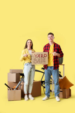 Photo for Young couple pointing at cardboard with text YARD SALE and boxes of unwanted stuff on yellow background - Royalty Free Image