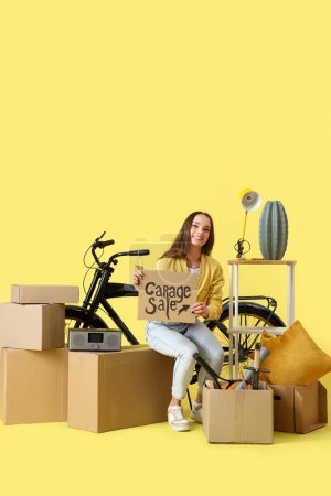 Photo for Young woman holding cardboard with text GARAGE SALE and boxes of unwanted stuff on yellow background - Royalty Free Image