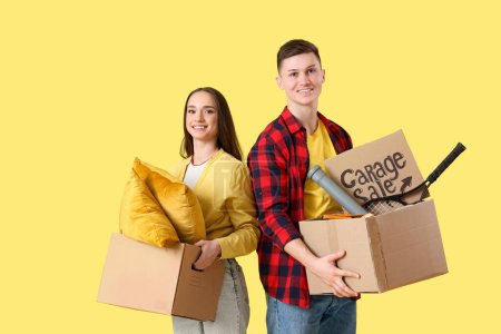 Photo for Young couple holding cardboard with text GARAGE SALE and boxes of unwanted stuff on yellow background - Royalty Free Image
