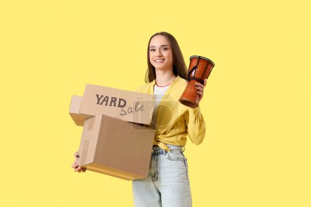 Photo for Young woman holding cardboard with text YARD SALE and boxes of unwanted stuff on yellow background - Royalty Free Image