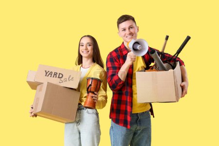 Photo for Young couple holding cardboard with text YARD SALE and boxes of unwanted stuff on yellow background - Royalty Free Image