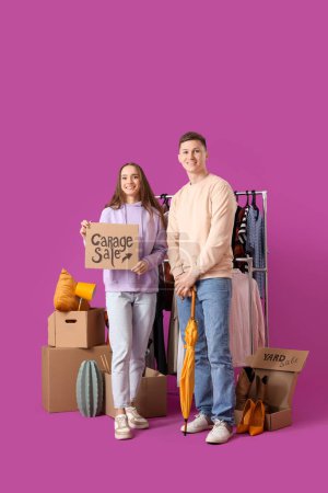Photo for Young couple holding cardboard with text GARAGE SALE and boxes of unwanted stuff on purple background - Royalty Free Image