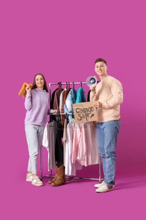 Photo for Young couple holding cardboard with text GARAGE SALE and unwanted clothes on purple background - Royalty Free Image