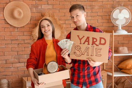 Photo for Young couple holding cardboard with text YARD SALE and money in room of unwanted stuff - Royalty Free Image