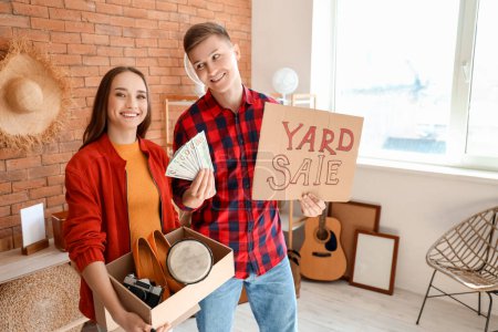 Photo for Young couple holding cardboard with text YARD SALE and money in room of unwanted stuff - Royalty Free Image