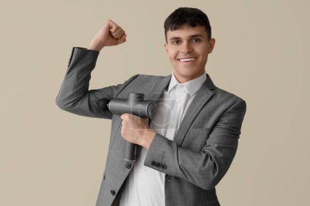 Junger Geschäftsmann massiert seinen Arm mit perkussivem Massagegerät auf beigem Hintergrund