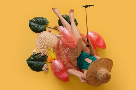 Belle jeune femme en maillot de bain avec anneau gonflable et feuilles de palmier prenant selfie sur fond jaune