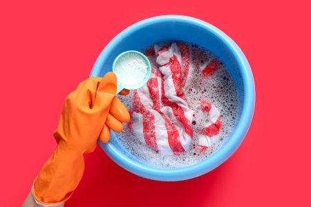 Woman in rubber gloves adding laundry detergent to clothes in plastic basin on red background