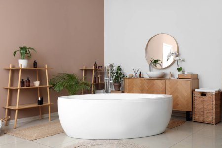 Interior of stylish bathroom with bathtub, houseplants and wooden furniture