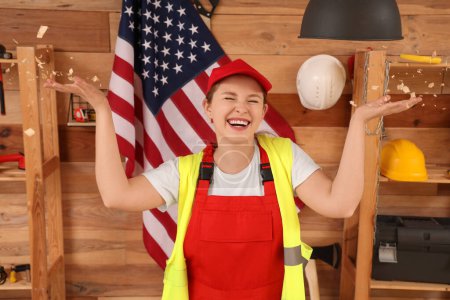 Young female carpenter with USA flag and sawdust having fun in workshop. Labor Day celebration