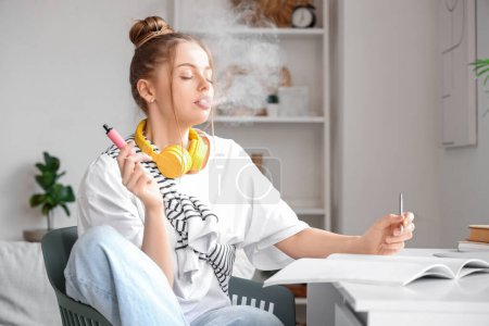 Téléchargez les photos : Jeune femme fumant une cigarette électronique jetable et écrivant sur un ordinateur portable à la maison - en image libre de droit
