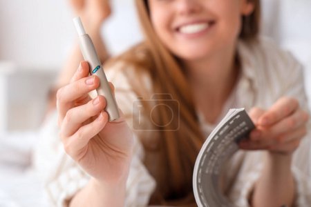 Téléchargez les photos : Jeune femme lisant le magazine et fumant le cigare électronique sur le lit à la maison, gros plan - en image libre de droit