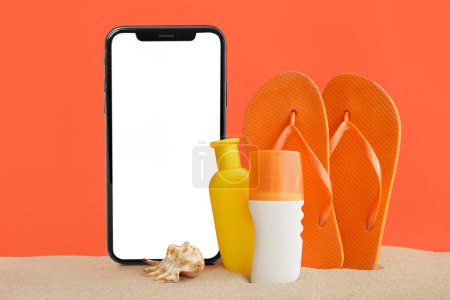 Foto de Gran teléfono móvil con pantalla vacía, protectores solares y chanclas sobre arena contra fondo naranja - Imagen libre de derechos