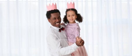 Photo for Happy African-American man and his little daughter in paper crowns dancing at home - Royalty Free Image