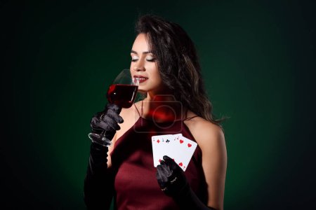 Beautiful young African-American woman with playing cards and glass of wine on dark green background