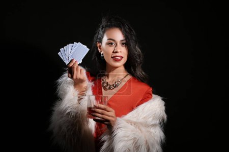 Beautiful young African-American woman with glass of whiskey and playing cards on black background