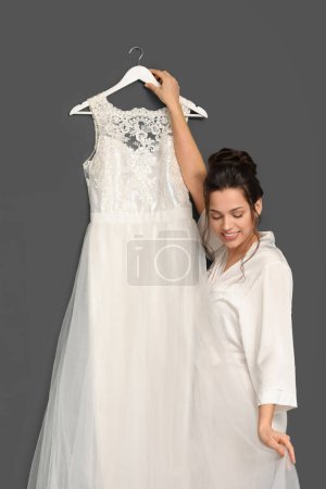 Foto de Hermosa novia joven con su vestido de novia colgando en la pared oscura - Imagen libre de derechos