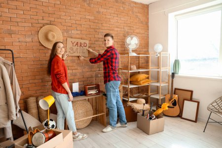 Photo for Young couple hanging cardboard with text YARD SALE on wall in room of unwanted stuff - Royalty Free Image