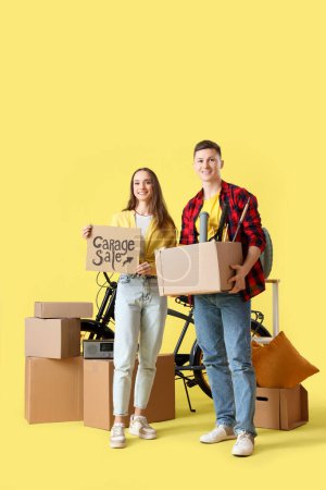 Photo for Young couple holding cardboard with text GARAGE SALE and boxes of unwanted stuff on yellow background - Royalty Free Image