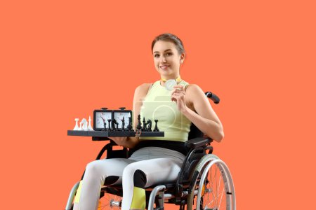 Young woman in wheelchair with first place medal and chess on orange background