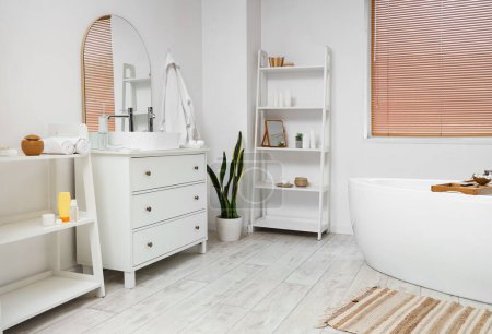 Interior of light bathroom with bathtub, chest of drawers and houseplant