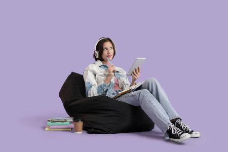 Photo for Female student in headphones with tablet computer and copybooks studying on beanbag against lilac background - Royalty Free Image