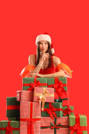 Young woman in Santa hat with swim armbands and gifts showing silence gesture on red background. Christmas in July