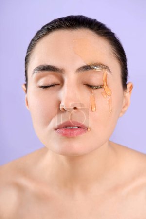Young woman with dripping honey on her face against lilac background, closeup