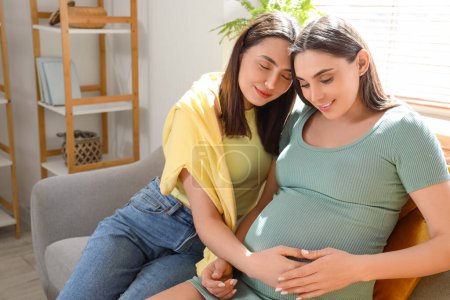 Young lesbian woman hugging her pregnant wife on sofa at home