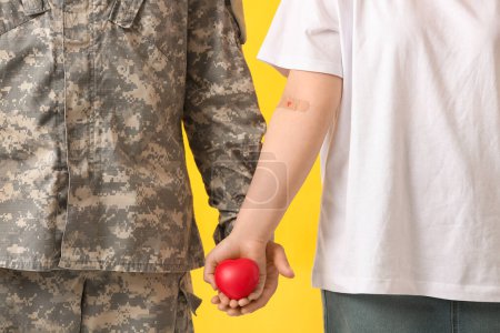 Blood donor with applied medical patch and soldier holding heart on yellow background