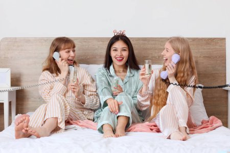 Young woman and her bridesmaids with champagne talking by phones at Hen Party in bedroom