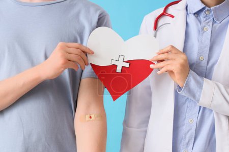 Male blood donor with doctor holding paper heart on blue background, closeup