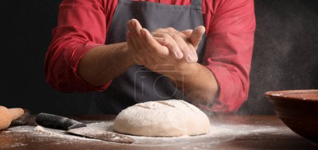Männlicher Bäcker streut Teig mit Mehl am Tisch auf dunklem Hintergrund