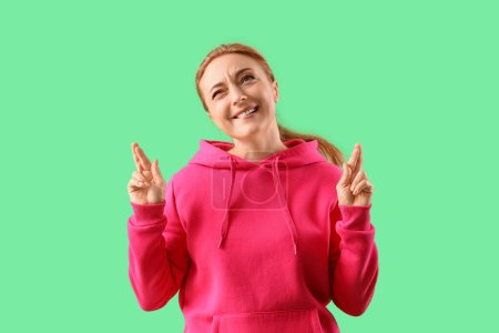 Mature woman in fuchsia hoodie crossing fingers on green background