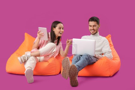 Beautiful young happy couple with laptop and tablet resting on beanbag chairs against purple background