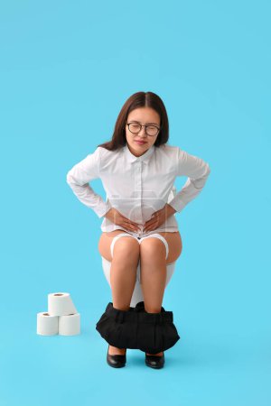 Téléchargez les photos : Jeune femme d'affaires asiatique avec mal de ventre assis sur un bol de toilette sur fond bleu - en image libre de droit