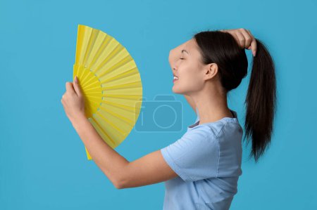 Téléchargez les photos : Jeune femme avec ventilateur de main souffrant de chaleur sur fond bleu - en image libre de droit