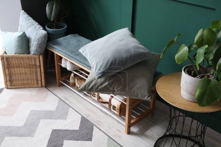 Gemütliche Bank, Schuhe, Kissen und Zimmerpflanze neben grüner Wand in schönem Raum
