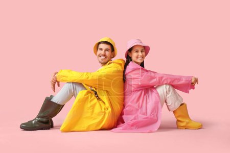 Young couple in raincoats sitting on pink background