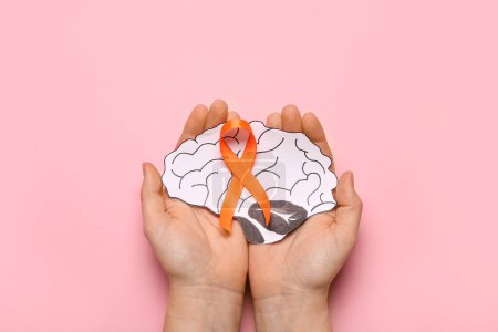Female hands with orange ribbon and paper brain on pink background. Multiple Sclerosis Awareness Month