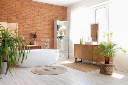 Interior of stylish bathroom with bathtub, sink, chest of drawers and houseplant