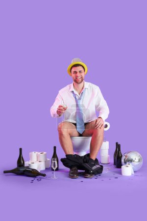 Téléchargez les photos : Jeune homme d'affaires ivre assis sur un bol de toilette après la fête sur fond lilas - en image libre de droit